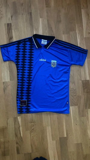 Argentina Retro 1994 - Retro 1994 Argentina fotbollströja från Adidas i blankt blått material med svarta och blå geometriska mönster längs sidan. Kortärmad med svart krage och vita detaljer. Broderat AFA-emblem på bröstet och Adidas-logga. Vid fler frågor/funderingar tveka inte på att skriva!