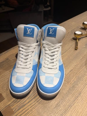 Louis Vuitton blåvita high-top sneakers - Säljer ett par Louis Vuitton high-top sneakers i blått och vitt med rutigt mönster. Skorna har snörning, rund tå och en bred rem med LV-logga bak. Tillverkade i skinn och textil med coola detaljer och tydlig branding på både plös och häl. Perfekt för dig som gillar statement pieces.