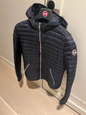 Mörkblå Colmar pufferjacka med huva - Snygg mörkblå pufferjacka från Colmar med huva och två dragkedjeförsedda fickor. Jackan har quiltad design och raka linjer, perfekt för dig som gillar stilren och sportig look. Märkeslogga på ärmen och hel dragkedja framtill. Passar tjej ca 160-165cm NYSKICK!!