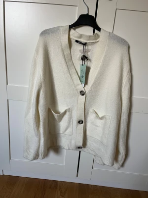 Vit stickad kofta från Gina Tricot - Supermysig vit stickad kofta från Gina Tricot med v-ringad hals, stora knappar och två rejäla fickor framtill. Perfekt för lager-på-lager och chill dagar. Ribbad kant nedtill och vid ärmslut för extra comfy vibe. Ny med prislapp, strl M.