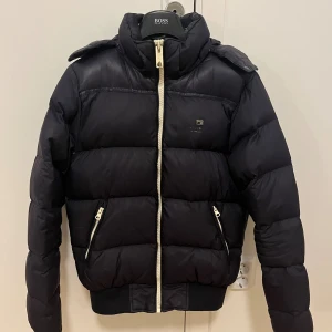 Mörkblå pufferjacka från Scotch & Soda - Svart pufferjacka från Scotch & Soda med hög krage och avtagbar huva. Jackan har vita dragkedjor framtill och på fickorna, samt ribbade muddar vid nederkant och ärmslut. Perfekt för kalla dagar med sin tjocka vaddering och stilrena look. Pris kan diskuteras!