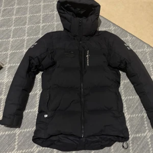 Svart dunjacka från Sail Racing - Svart dunjacka från Sail Racing med huva och dragkedja framtill. Jackan är vadderad med 500 FP dun från Allied Feather + Down och har en stilren look med logga på bröstet och ärmen. Perfekt för kalla dagar och har en längre passform baktill.