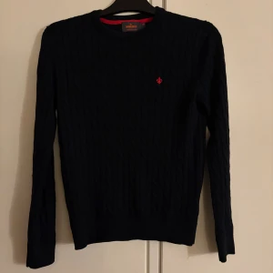 Mörkblå kabelstickad tröja Morris - Kabelstickad mörkblå tröja från Morris i 100% merinoull. Klassisk rund hals och långärmad modell med röd broderad logga på bröstet. Perfekt för dig som gillar stilrena och tidlösa plagg med snygga detaljer. Sparsamt använd, skick: 9/10. Nypris på hemsidan: 1699kr, Mitt pris: 399kr