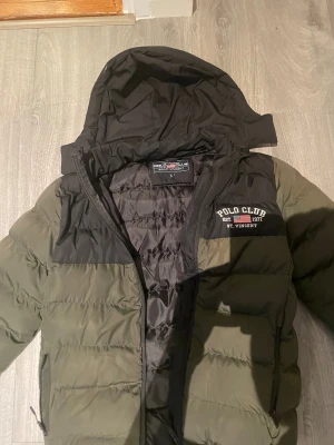 Grön pufferjacka Polo Club S - Snygg pufferjacka från Polo Club St. Vincent i olivgrönt med svarta detaljer och huva. Jackan har broderad logga på bröstet, dragkedja framtill och två sidofickor. Perfekt för kyliga dagar och riktigt skön att bära.