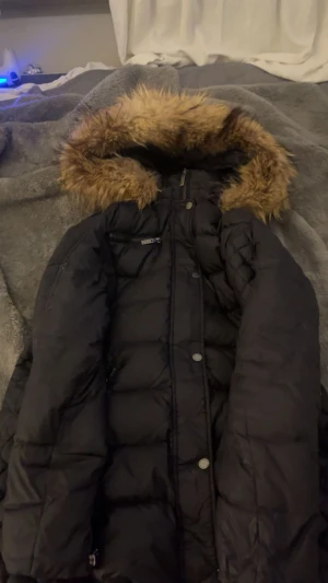 rocknblue  - Säljer en svart dunjacka med fluffig fuskpäls runt huvan. Jackan har långa ärmar, dragkedja och knappar framtill. Den är quiltad och har en snygg, lite längre passform som håller dig varm under kalla dagar. Perfekt för dig som gillar stilren och cozy vinterlook.