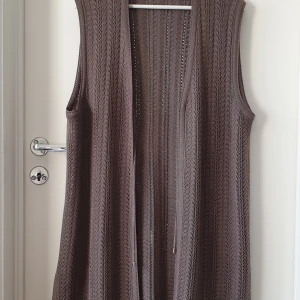 Taupe stickad lång väst med snörning - Säljer en brun, lång och stickad väst med snyggt hålmönster och smala snören framtill. Västen är ärmlös och har en loose passform, perfekt att slänga över en t-shirt eller skjorta för en chill look. 100% ak silk 