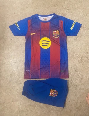 FC Barcelona Lamine Yamal 10 matchtröja - Säljer en FC Barcelona matchtröja med Lamine Yamal och nummer 10 på ryggen. Tröjan är kortärmad med klassiska blå och röda vertikala ränder, gula detaljer och klubbmärke på bröstet. Materialet är lätt och andas, perfekt för fotboll. Shorts med klubbmärke ingår.