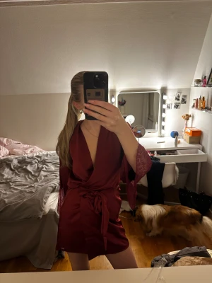 Vinröd morgonrock från hunkemöller - Supersnygg vinröd satin morgonrock från Noir by Hunkemöller med omlottmodell, knyt i midjan och spetsdetaljer på ärmar och rygg. Den har djup v-ringning och är tillverkad i mjuk satinliknande polyester. 