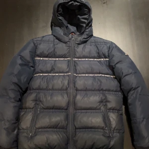 Mörkblå pufferjacka Tommy Hilfiger - Snygg mörkblå pufferjacka från Tommy Hilfiger som ej säljs längre! Jackan är i ett bra skick, lite smutsig med någon enstaka repa. Storlek 176, passar dig mellan 170-178 skulle jag bedöma! Skriv vid frågor! 