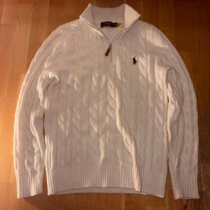 Vit kabelstickad tröja Polo Ralph Lauren - Vit kabelstickad tröja från Polo Ralph Lauren med half zip och brunt läderdetalj på dragkedjan. Tröjan har ribbad krage, muddar och nederkant samt det klassiska broderade logot på bröstet. Perfekt för dig som gillar preppy stil och snygga detaljer.