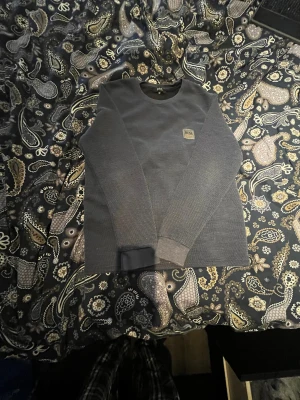 Mörkgrå långärmad tröja Hugo Boss - Snygg mörkgrå långärmad tröja från Hugo Boss med diskret rutmönstrad struktur och rund hals. Tröjan har en liten patch med BOSS-loggan på bröstet och ribbade muddar vid ärmslut och nederkant. Perfekt för dig som gillar stilrena och klassiska plagg.