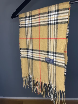 Burberry Halsduk Beige - Klassisk rutig halsduk från Burberry i 100% lammull. Beige bas med svarta, vita och röda rutor samt fransar i ändarna. Perfekt accessoar för att lyfta din outfit med ikoniskt brittiskt mönster.