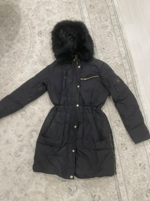 Svart parkas från Hollies med äkta päls. - Svart parkas från Hollies med snygga gulddetaljer och dragkedjor. Jackan har en stor huva med fluffig äkta päls, markerad midja och flera fickor. Perfekt för kalla dagar och riktigt stilren look.