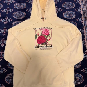 Gul hoodie med rosor från HUF - Snygg gul hoodie från HUF med tryck av röda och rosa rosor samt texten 'Destroy & Rebuild' och 'huf worldwide' på bröstet. Tröjan har huva med snörning och stor magficka. Perfekt för dig som gillar streetwear och coola prints.