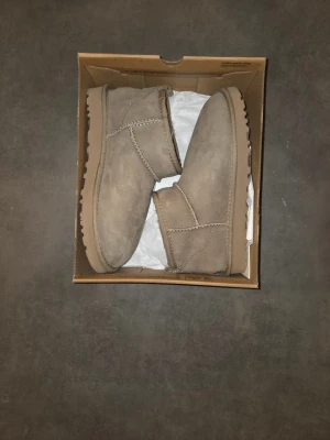 Beige UGG boots i mocka - Säljer ett par klassiska beige UGG boots i mocka med låg skaft och mjukt foder. Skorna har rund tå, platt sula och dragflik bak för enkel påtagning. Perfekta för kyliga dagar.