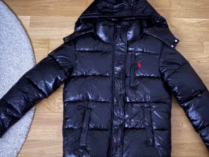 Svart pufferjacka från Polo Ralph Lauren - Säljer en svart pufferjacka från Polo Ralph Lauren med glansig finish och röd broderad logga på bröstet. Jackan har huva, dragkedja framtill och flera fickor. Perfekt för kalla dagar. Går gärna ner i pris vid snabb affär.