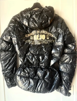 Dun pufferjacka från Kejo med brodyr - Snygg svart dun pufferjacka från Kejo med glansig finish, broderade detaljer i guld på ryggen och ärmen. Jackan har huva, dragkedja framtill och markerad midja. Perfekt för dig som vill sticka ut med en unik och trendig vinterjacka.