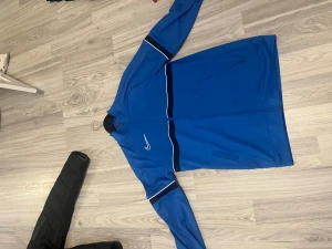 Blå Nike vindjacka Dri-Fit M - Snygg blå vindjacka från Nike med Dri-Fit-teknologi. Jackan har hel dragkedja, vita och svarta detaljer på bröstet och ärmarna samt två fickor med dragkedja. Perfekt för dig som gillar sportig stil och vill ha något lätt och funktionellt    MÖTS I STOCKHOLM🙃
