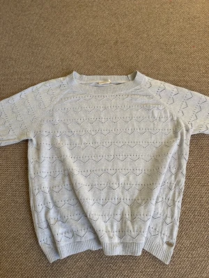 Ljusgrå stickad tröja från Dobber - Snygg ljusgrå stickad tröja från Dobber med hjärtmönster och ribbade muddar vid hals, ärmslut och nederkant. Tröjan har lång ärm och en liten metallbricka med Dobber-logga vid nederkanten. Perfekt för dig som gillar mjuka och stilrena plagg.