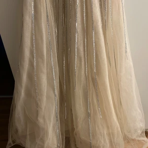 Champagnefärgad långklänning med glitter - En magisk champagnefärgad långklänning med tunna axelband och djup v-ringning. Klänningen har glittriga silverränder som ger en lyxig känsla och ett skirt, svepande tyg som faller snyggt. Perfekt för bal eller nyår, du kommer definitivt sticka ut i denna drömklänning!