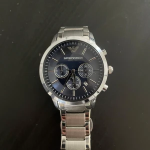 Emporio Armani silverfärgad klocka - Stilren klocka från Emporio Armani med mörkblå urtavla och silverfärgat armband i metall. Klockan har tre mindre urtavlor, datumvisning och tydliga indexmarkeringar. Klassisk rund form och robust länkarmband ger en lyxig känsla.
