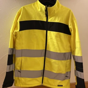 Reflex vindjacka från Bluewear - Gul och svart vindjacka från Bluewear med reflekterande gråa ränder. Jackan har hel dragkedja framtill, hög krage och två fickor. Perfekt för dig som vill synas och hålla dig skyddad mot vinden. Snygg och funktionell design. Smutsen går nog bort i tvätten men har inte testat. Annars snus det knappt att den är använd.