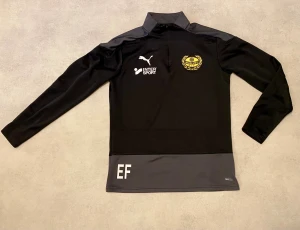 Svart Puma Mjällby AIF half zip träningströja - Svart långärmad träningströja från Puma med Mjällby AIF-logga i gult på bröstet och sponsormärken på ryggen. Tröjan har grå nederkant, half zip vid kragen och är gjord i polyester med dryCELL-teknologi. Perfekt för fotbollsträning och sportiga dagar.
