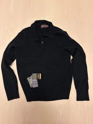 Mörkblå stickad kragtröja från Morris - Mörkblå tröja från Morris med klassisk krage och knappar framtill. Tröjan är tillverkad i Merinoull och har långa ärmar samt ribbade muddar vid ärmslut och nederkant. Perfekt för dig som gillar stilrena och tidlösa plagg.