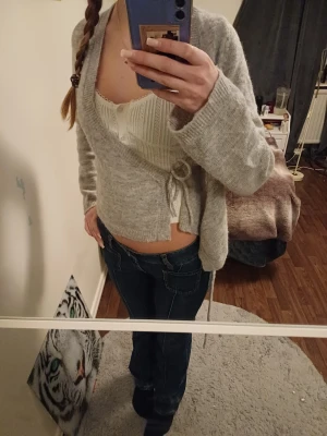 Ljusgrå stikad hudde med knyt - Säljer en ljusgrå stickad huddie med knyt i sidan. Hudden har v-ringad hals. vilket ger en snygg look. Perfekt att slänga över en topp för extra lager. 