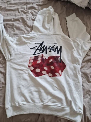 Vit Stüssy hoodie med tärningsprint - Vit hoodie från Stüssy med svart logga på bröstet och stort tryck av röda tärningar och logga på ryggen. Klassisk känguruficka och huva. Perfekt för dig som gillar streetwear och vill sticka ut med en unik design.