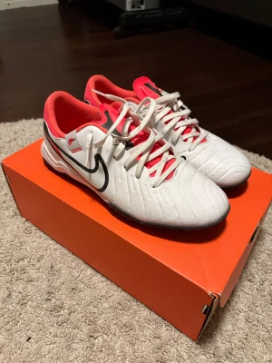 Nike Tiempo vita fotbollsskor - Nike Tiempo fotbollsskor i vitt med svarta detaljer och inslag av rött. Skorna har klassisk snörning, låg profil och en ovandel i syntetmaterial. Swoosh-logga på sidan och röd insida för extra pop. Perfekta för dig som vill sticka ut på planen.