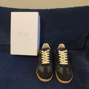 Maison Margiela svarta sneakers strl 44 - Maison Margiela sneakers i svart skinn och mocka med beige gummisula och vita snören. Klassisk rund tå och platt sula, stilren design med kontrasterande material och subtil branding på sulan. Perfekt för dig som gillar minimalistisk och lyxig streetstil.
