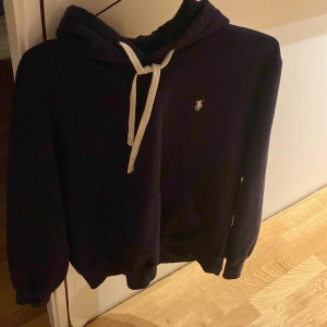 Mörkblå hoodie från Polo Ralph Lauren - Snygg mörkblå hoodie från Polo Ralph Lauren med vit snörning och klassisk logga broderad på bröstet. Tröjan har huva, magficka och ribbade muddar vid ärmslut och nederkant. Perfekt för chill dagar och enkel att matcha med jeans eller joggers.
