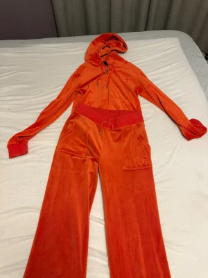 Juicy Couture orange hoodie i velour - Juicy Couture hoodie i stark orange velour med huva och dragkedja framtill. Långärmad med breda muddar och snörning vid halsen. Ikonisk JC-logga broderad på bröstet. Perfekt för dig som gillar retro vibes och comfy stil.