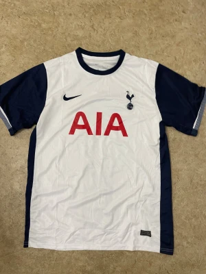 Tottenham Hotspur vit/blå Nike tröja - Säljer en Tottenham Hotspur fotbollströja från Nike med AIA sponsortryck på bröstet. Tröjan är vit med mörkblå ärmar och detaljer, har rund hals och korta ärmar. Materialet är lätt och andas, perfekt för match eller träning. Klubbmärket sitter på vänster bröst.