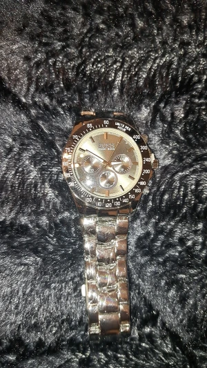 Silverfärgad Hugo Boss kronografklocka - Säljer en stilren kronografklocka från Hugo Boss i silverfärgat metall med länkarmband. Urtavlan har tre mindre visare och markerade index, samt svart ring med vita siffror runt boetten. Boss-logga på både urtavla och spänne. Perfekt för dig som gillar klassisk design. Helt ny med plastet på