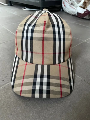 Burberry rutig keps i beige och svart - Snygg keps från Burberry med klassiskt rutigt mönster i beige, svart, vitt och röda detaljer. Märke med 'Burberry London England' baktill. Kepsen har böjd skärm och justerbar rem. Perfekt för dig som gillar ikonisk design och vill sticka ut.