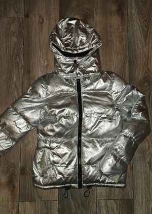 Silvrig pufferjacka med huva - Säljer en riktigt fet silvrig pufferjacka med huva och svart dragkedja. Jackan har quiltad look, dragsko nertill och stora fickor. Perfekt för dig som vill sticka ut i vinterkylan.