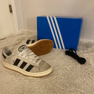 Adidas Campus 00s W beige/svarta sneakers - Adidas Campus 00s W sneakers i beige mocka med svarta ränder och vit sula. Klassisk retrostil med rund tå, platt gummisula och tydlig Adidas-logga på plösen. Skorna har vita skosnören och extra svarta skosnören medföljer. Perfekta för dig som gillar streetwear.