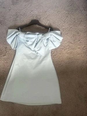 Ljusblå offshoulder kortklänning - Supersnygg ljusblå kortklänning med offshoulder-design och stora volangdetaljer över axlarna. Klänningen har en figurnära passform och är tillverkad i ett slätt, mjukt material som ger en elegant känsla. Perfekt för dig som vill sticka ut med en unik look.