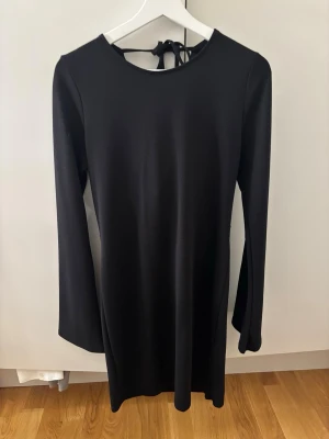 Svart klänning med öppen rygg från Lager 157 - Säljer en svart, stilren klänning från Lager 157 i storlek M. Klänningen har långa ärmar, kort längd och en snygg öppen rygg med knytband i nacken. Materialet känns mjukt och följsamt, perfekt för dig som gillar minimalistisk design med en twist.