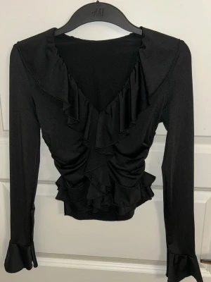 Svart volangblus från H&M - Snygg svart blus med v-ringning och volangdetaljer längs halsringningen och nederkanten. Långärmad modell med lätt figurnära passform och volang vid ärmslut. Perfekt att styla med jeans eller kjol för en trendig look. Använt 1 gång 