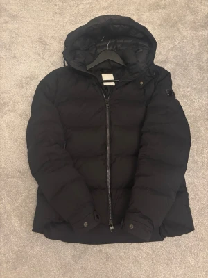 Svart pufferjacka från J.Lindeberg - Säljer en svart pufferjacka från J.Lindeberg med huva och dragkedja framtill. Storlek L och man bör vara runt 185 och uppåt för att jackan ska sitta bra. Jackan har en stilren look med diskret logga på ärmen och bra material som håller dig varm. Perfekt för kalla dagar och enkel att matcha med allt. Skriv vid minsta fråga eller fundering!