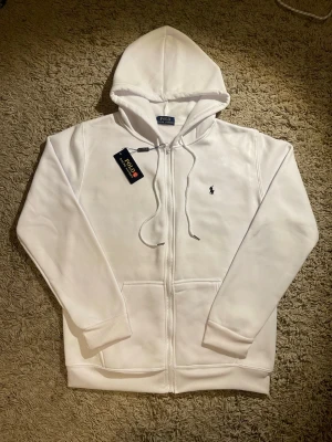 Vit hoodie från Polo Ralph Lauren - Vit hoodie från Polo Ralph Lauren med klassisk svart logobrodyr på bröstet. Tröjan har huva med snörning, dragkedja framtill och två Vit hoodie från Polo Ralph Lauren fickor. Materialet är mjukt och känns skönt mot huden, perfekt för chill dagar. Snygg och enkel design som alltid funkar.