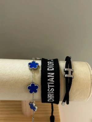 3st armband i blå och svart färg - Säljer en blå väst från Christian Dior med broderad svart text och vita detaljer. Västen har en klassisk rak form och är tillverkad i ett syntetmaterial. Ett perfekt statement-plagg för dig som vill sticka ut med en ikonisk och elegant design.  ✨ Specialerbjudande: Tre armband säljs tillsammans för endast 200 kr.