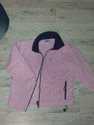 Rosa fleecejacka från Nike - Mysig rosa fleecejacka till barn Nike med svart krage och hel dragkedja framtill. Jackan har två fickor med dragkedja och en liten Nike-logga på bröstet. Perfekt för kyliga dagar och enkel att slänga på sig över en hoodie.