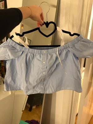Ljusblå offshoulder topp med knappar - Säljer en ljusblå croppad offshoulder topp med vita knappar framtill och vita band som axelband från pull&bear. Toppen har puffiga korta ärmar och är gjord i ett tunt bomullsliknande material. Perfekt för varma dagar och en söt, avslappnad stil.