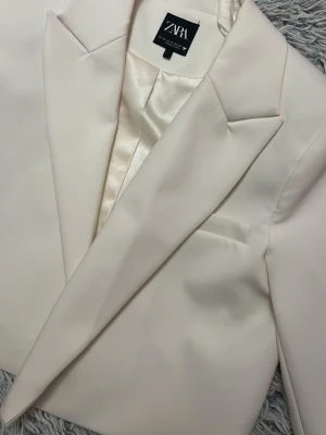 Vit croppad kavaj från Zara - Säljer en stilren vit croppad kavaj från Zara med klassisk krage och slät finish. Kavajen har lång ärm och är fodrad med glansigt tyg. Perfekt för dig som vill ha en clean och modern look.