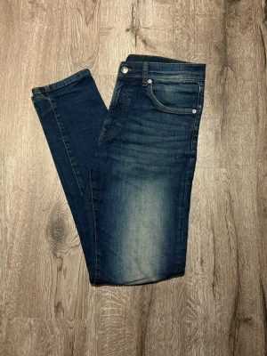 Slim fit jeans från East West - Mörkblå slim fit jeans från East West med snygg tvätt och klassiska fem fickor. Jeansen har en lätt slitning framtill och bak, normal midja och raka ben. Perfekta för dig som gillar en smalare passform och stilren look. Storlek 30/32! 