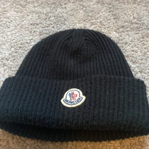 Svart Moncler mössa i ull - Svart ribbstickad mössa från Moncler med klassisk uppvikt kant och broderad logotyp framtill. Tillverkad i mjuk ull som håller dig varm under kyliga dagar. Perfekt accessoar för att lyfta din vinterstil.